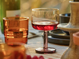 A glass of red wine sits on a table set with brown glassware, black dishes, and a beige tablecloth in natural light.