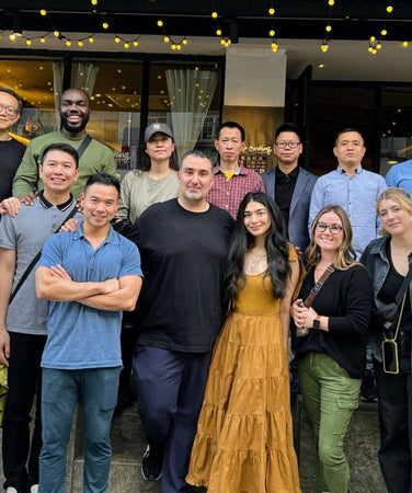 A group of sixteen people pose together outside a restaurant, standing and kneeling in two rows, with string lights overhead.