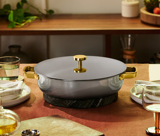 A gray pot with gold handles and lid sits on a dark trivet in the center of a set dining table with dishes, glasses, and napkins.