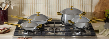 Four stainless steel pots and pans with gold handles are arranged on a gas stove, surrounded by kitchen utensils, vegetables, and a plant.