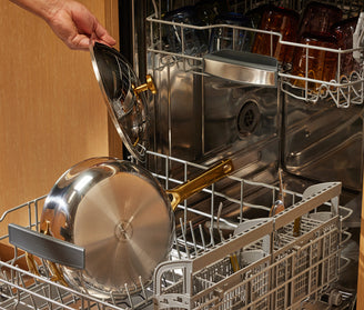 A person places a stainless steel pan and its lid into the bottom rack of a dishwasher filled with other dishes.