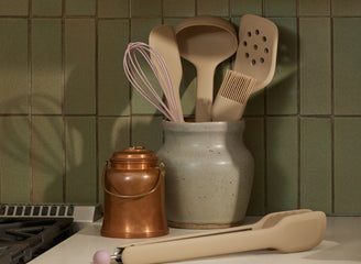 Set of kitchen utensils including a whisk, ladle, and spatula in a ceramic holder against a green tiled wall.