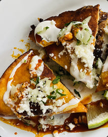 Two slices of birria quesadilla topped with crema, crumbled cheese, onions, and cilantro on a white plate with a lime wedge and drizzled sauce.