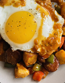 A fried egg topped with sauce sits on a bed of roasted potatoes, sausage slices, and diced bell peppers.