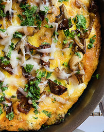 Close-up of a baked egg frittata in a skillet topped with melted cheese, mushrooms, and chopped fresh herbs.