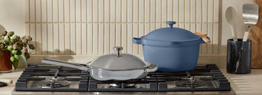 Blue cast ceramic pot and titanium pan on a stove with kitchen utensils in the background.