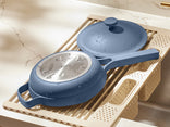 A blue nonstick frying pan and matching lid with water droplets rest on a beige dish rack above a kitchen sink.