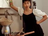 A person in a black dress with white puffed sleeves stands in a kitchen beside a stove with a pan, utensils, a woven hat, and a bottle of oil visible.