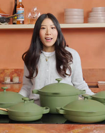 A woman stands in a kitchen behind a set of green cookware placed on a countertop with shelves of dishes and bottles in the background.