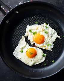 Two sunny-side up eggs sprinkled with black pepper and herbs in a non-stick frying pan.
