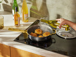 A person pours a light-colored liquid from a bottle into a frying pan with four pieces of chicken cooking on an induction stove.
