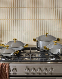 Four stainless steel pots with gold handles and lids are arranged on a gas stove, with a brown checkered towel hanging on the oven handle. Cream-colored tiles are on the wall behind.