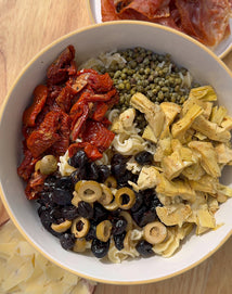 A bowl of pasta topped with sun-dried tomatoes, capers, artichoke hearts, black and green olives, next to a plate of sliced cured meat and cheese.