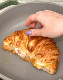 A hand with purple nail polish holds a croissant filled with cheese and ham on a grey nonstick pan.