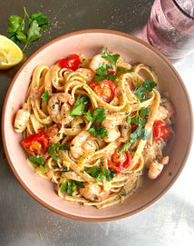A bowl of creamy shrimp and tomato pasta garnished with parsley, placed on a metal surface next to a lemon wedge and a glass of water.