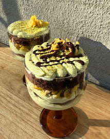 Two glass dessert cups filled with layered matcha cream, chocolate cake, and crumbled toppings, with one topped with chocolate drizzle, on a wooden surface.