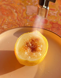 A lemon half is being caramelized with a kitchen torch on a beige plate, with a light brown and orange background.