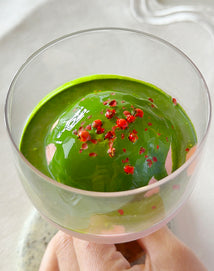 A hand holds a glass containing a green matcha dessert topped with pink peppercorns, set on a white surface.