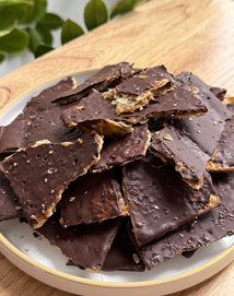 A plate filled with pieces of chocolate-covered cracker toffee, topped with sea salt, on a wooden surface with green leaves in the background.