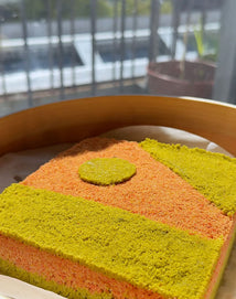 A square, colorful layered cake with orange and green sections and a round green piece on top, placed in a wooden container near a window with sunlight streaming in.