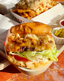 Close-up of a cheeseburger with lettuce, tomato, grilled onions, and sauce on a bun, with a side of fries topped with sauce and a small cup of pickles in the background.