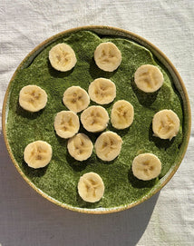 A bowl of green smoothie topped with evenly spaced banana slices, placed on a light-colored fabric surface.