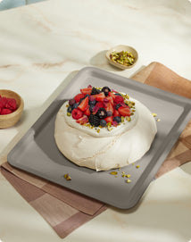 A pavlova topped with assorted fresh berries and chopped pistachios sits on a gray tray, with bowls of raspberries and pistachios nearby.