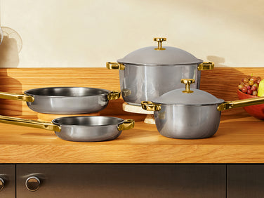 A set of stainless steel cookware with a matte gray finish and gold-tone handles and lids, arranged on a wooden table with a red runner in a kitchen setting.