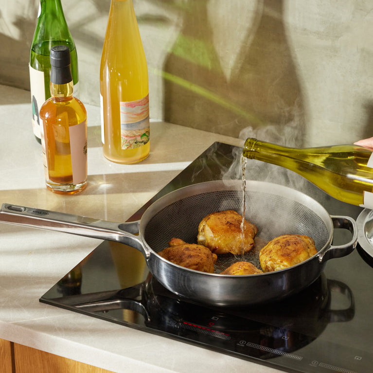 A hand pours white wine from a bottle into a frying pan with browned chicken pieces on an induction cooktop, with bottles on the kitchen counter nearby.