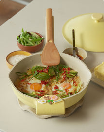 Yellow frying pan with food on a table, surrounded by ingredients and a wooden spatula.