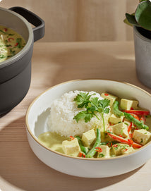 A bowl of white rice served with tofu and vegetable green curry, garnished with cilantro, sits on a wooden table next to a pot and a potted plant.