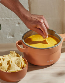 A hand dips a tortilla chip into a pot of cheese dip, with a bowl of chips beside the pot on a light wooden surface.