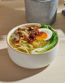 A bowl of ramen with noodles, bok choy, mushrooms, a halved soft-boiled egg, scallions, and chili oil, on a wooden table.
