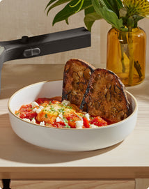 A white bowl with toasted bread slices, tomato-based stew, and crumbled cheese sits on a light wooden surface next to a yellow vase with green foliage.