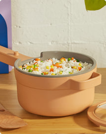 A beige saucepan filled with cooked rice and mixed vegetables sits on a wooden surface next to a spatula and its lid.