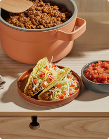 Two hard-shell tacos filled with ground meat, shredded lettuce, chopped tomatoes, and cheese on a plate, with a bowl of salsa and a pot of taco filling in the background.