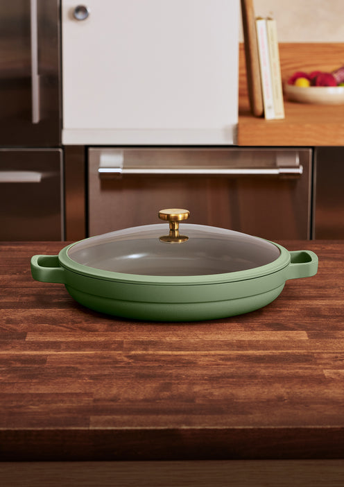 A green, nonstick skillet with a lid and two handles sits on a wooden kitchen counter, with modern cabinets and a fruit bowl in the background.