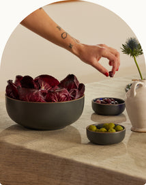 A hand reaches for a bowl of dark olives on a table set with a bowl of radicchio, a bowl of green olives, and a white ceramic pitcher.