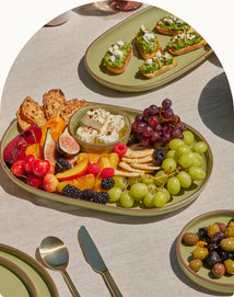 A green platter with assorted fresh fruits, crackers, and cheese sits on a beige tablecloth, with another plate of toasts and a small bowl of olives nearby.