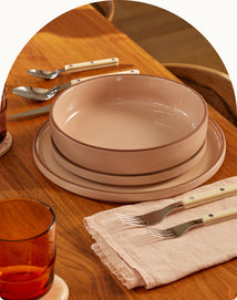 A pink plate and bowl are stacked on a wooden dining table set with cutlery, a pink napkin, and an amber glass.