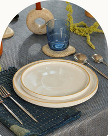 A set table with two stacked plates, a blue glass, two spoons, and a fork and knife on a blue textured napkin, atop a gray tablecloth.