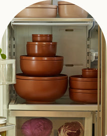Several stacked brown nesting bowls are arranged on a refrigerator shelf, with cabbages visible in the crisper drawer below.