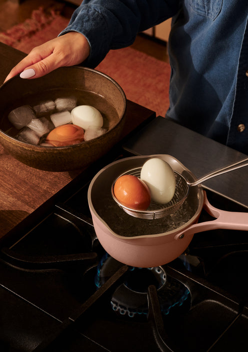 Person transferring boiled eggs from a saucepan of hot water on a stove into a bowl of ice water using a spoon.