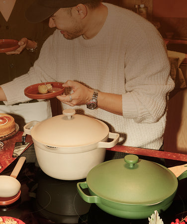 Two ceramic pots on a table with a person preparing food in the background.