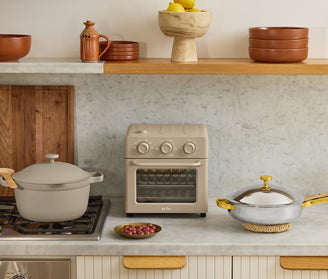 A beige countertop oven sits on a marble countertop between a covered pot on a stovetop and a lidded pan, with a small plate of grapes in front. Shelves hold bowls and a fruit stand.