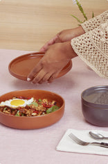 A person places a lid on a round brown food container filled with a meal topped with a fried egg, next to another closed container, utensils, and a napkin on a pink tablecloth.