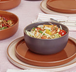 A ceramic bowl filled with mixed salad sits on stacked plates, surrounded by more bowls and plates on a pink tablecloth.