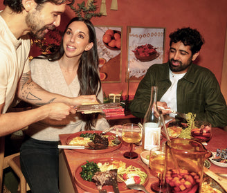 Three people sit around a table with plates of food and drinks, while one person grates cheese over a dish. The scene appears casual and friendly in a warmly lit room.