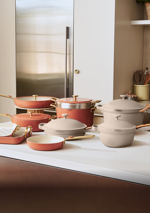 A collection of ceramic pots and pans in pink and beige colors with gold handles is displayed on a white kitchen counter.