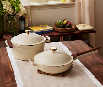 A beige saucepan with a lid and a beige skillet with a lid and wooden handles are placed on a wooden table with a runner in a kitchen setting.